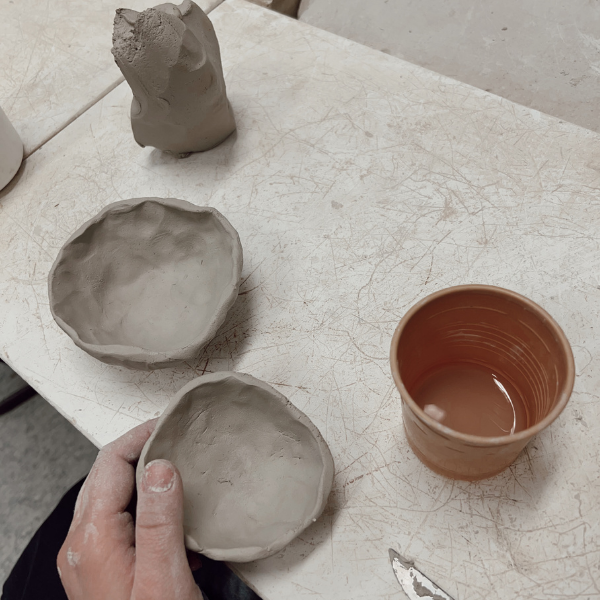 A hand holds a small handmade clay bowl on a light worktable, alongside two other unglazed clay bowls, a sculptural clay form, and a cup of water.