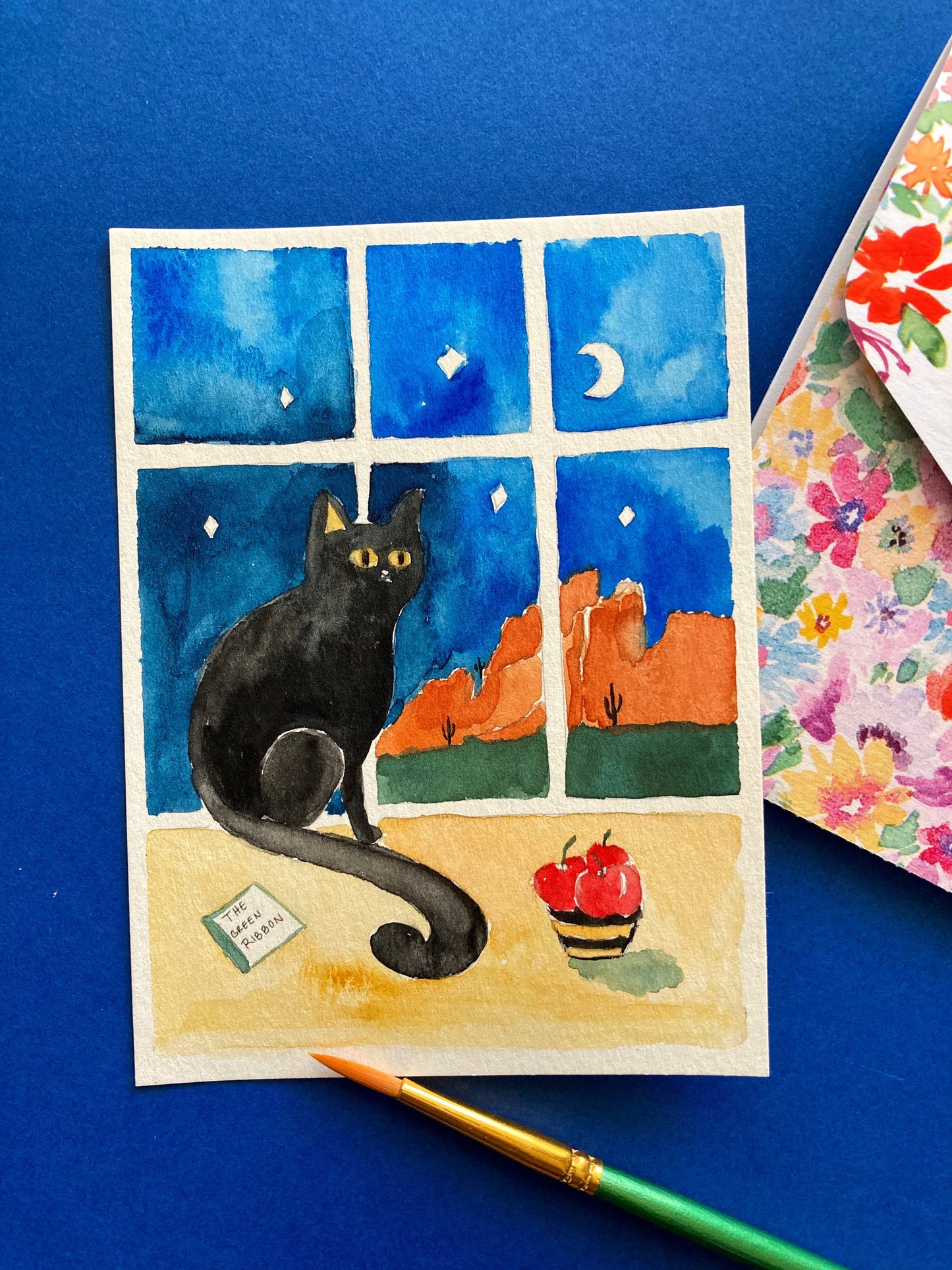 Watercolor painting of a black cat by a window with a blue sky and mountains, on a blue background.