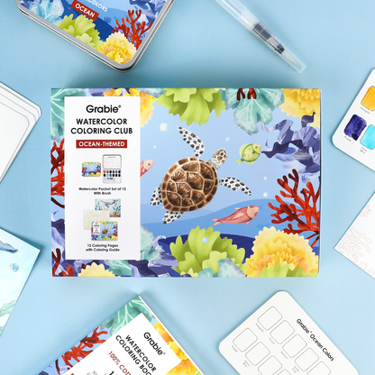 Grabie Watercolor Coloring Club Ocean-themed coloring book on a blue surface with art supplies.