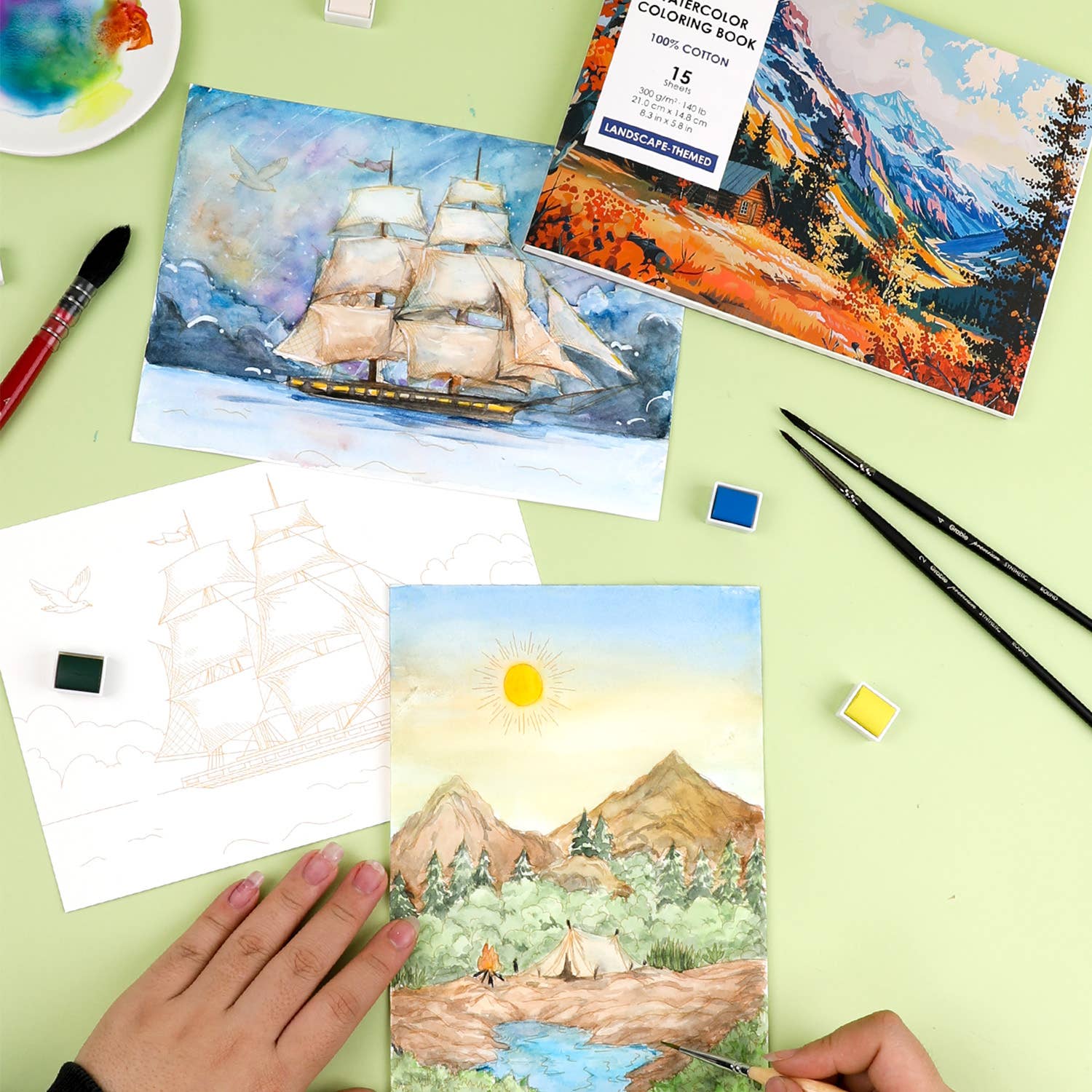 Coloring book with landscape designs on a green surface with art supplies.