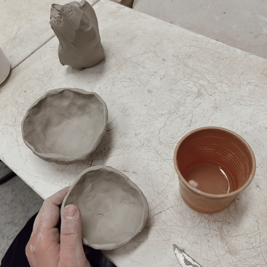 A hand holds a small handmade clay bowl on a light worktable, alongside two other unglazed clay bowls, a sculptural clay form, and a cup of water.