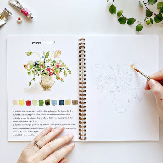 Artistic book with a painting of a poppy bouquet and color swatches, with a hand holding a paintbrush.