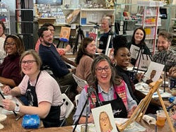 Group of people sitting around tables in a room with bookshelves and artwork.