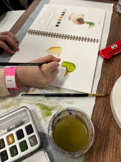 Person painting a leaf with watercolors on a sketchbook, with a watercolor set and cup of water nearby.