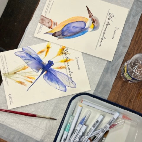 Art supplies including watercolor brushes, a dragonfly painting, and a notebook on a wooden surface.
